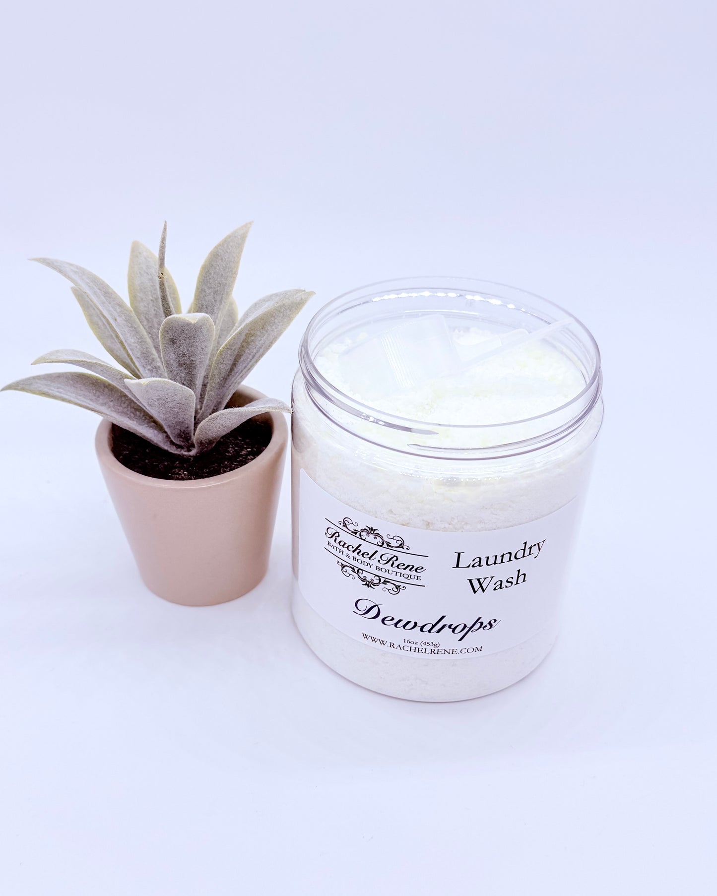 Jar of laundry wash labeled 'Dewdrops' next to a small potted plant on a white background