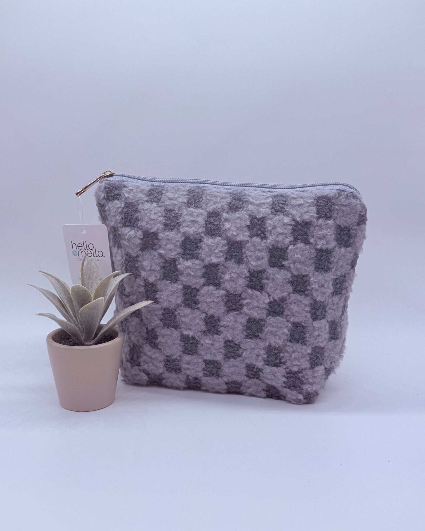 Gray checkered fuzzy cosmetic back with gold zipper top with a plant on a white background