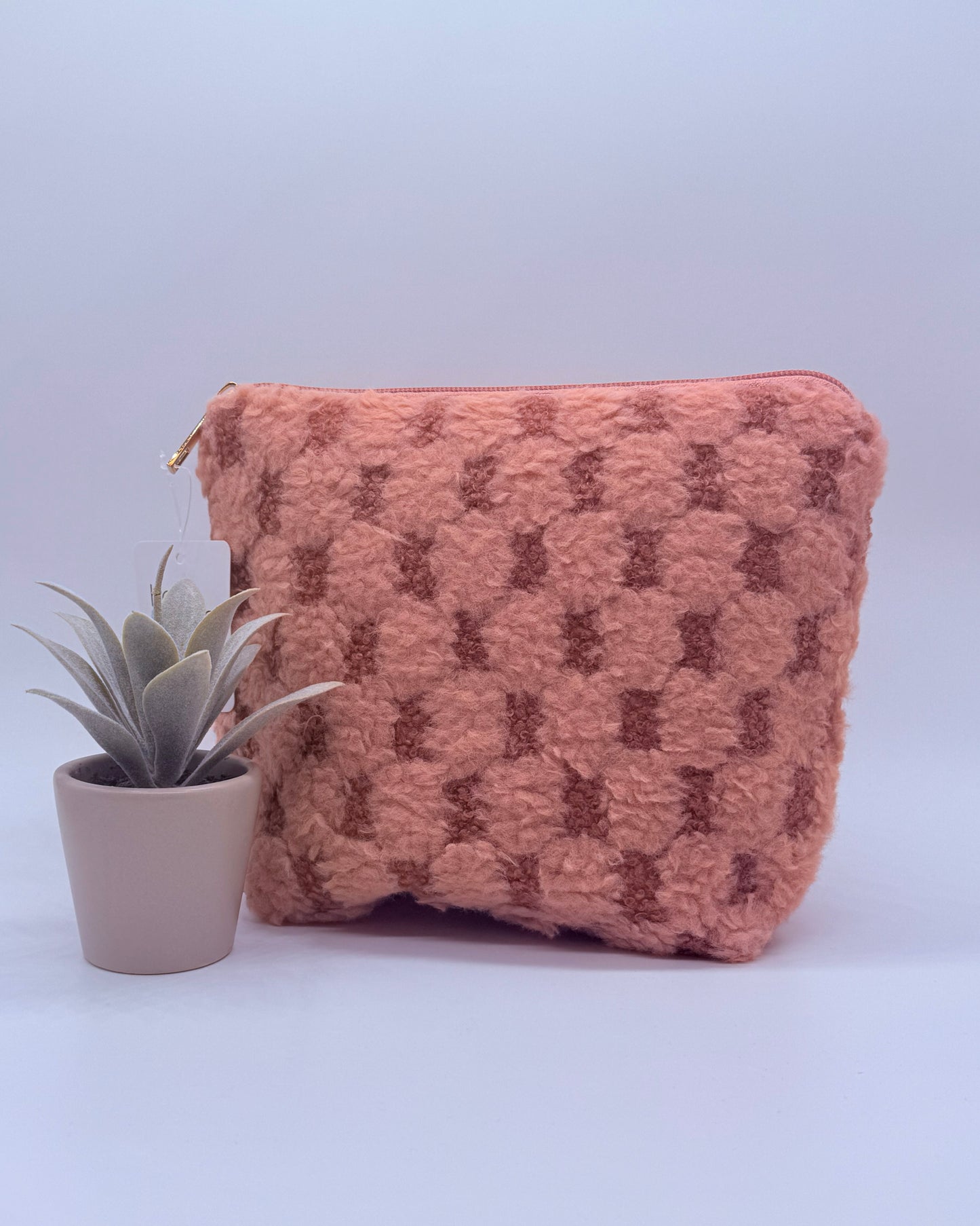 Pink checkered fuzzy cosmetic back with gold zipper top with a plant on a white background