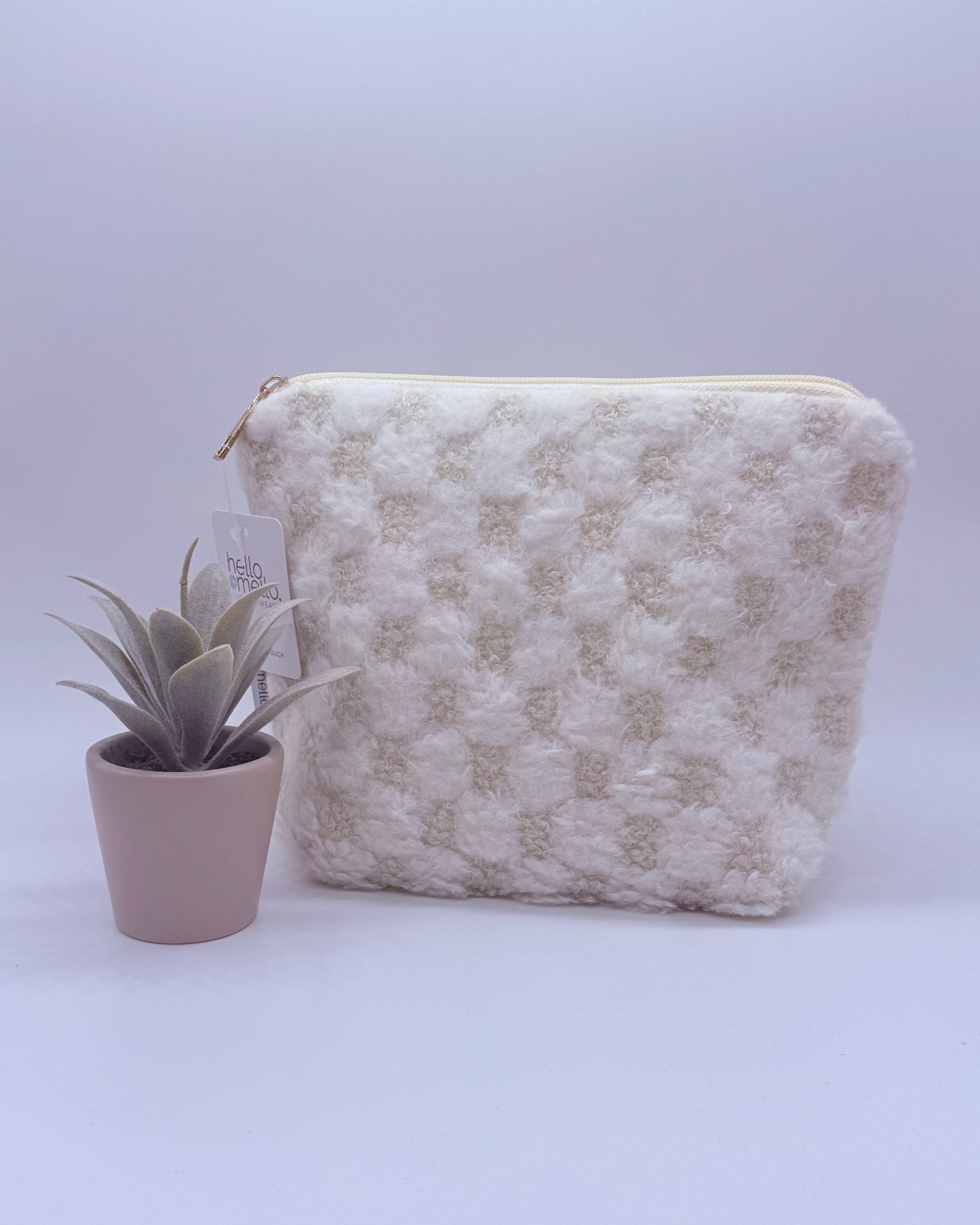 Cream colored checkered fuzzy cosmetic back with gold zipper top with a plant on a white background