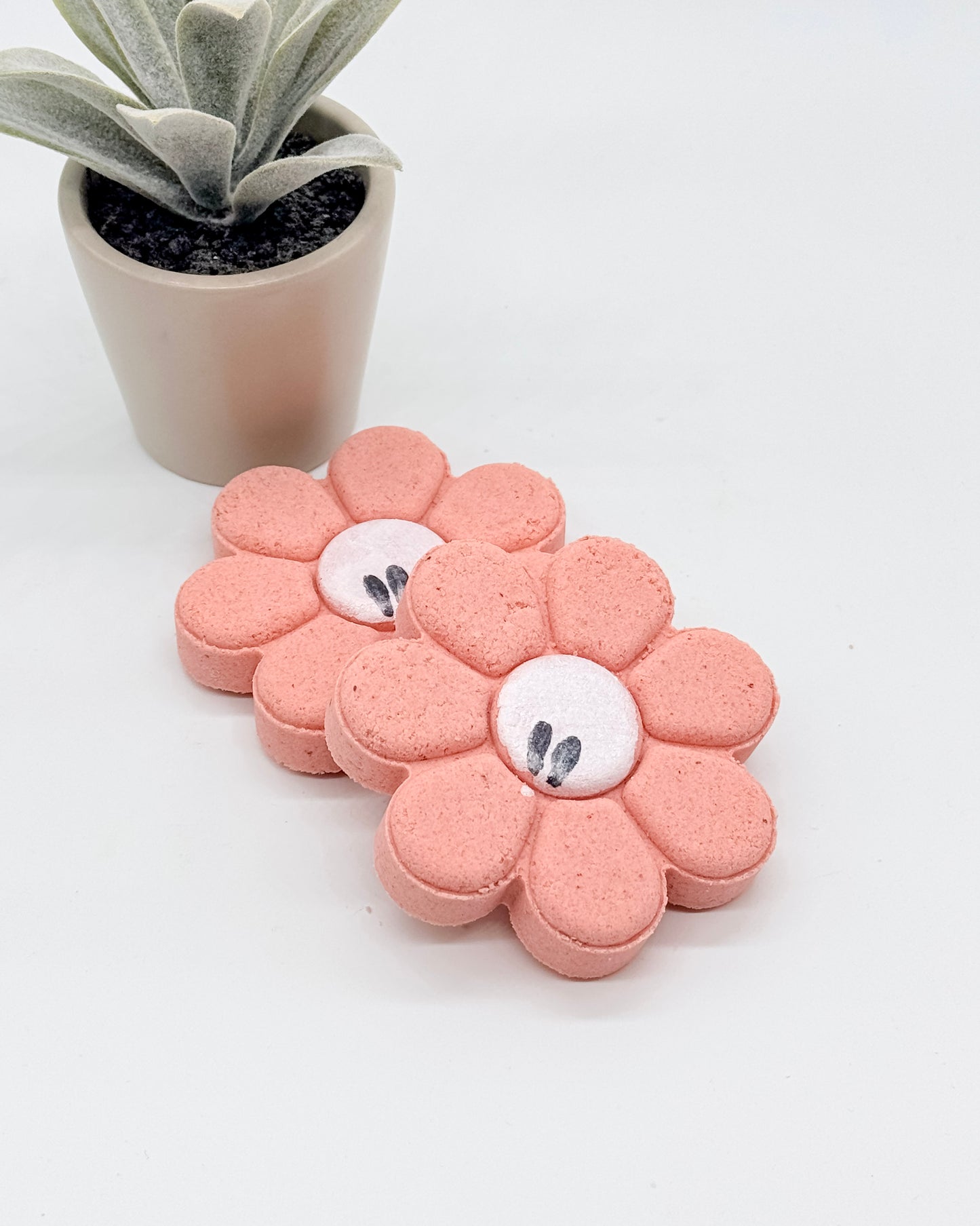 Pink flower-shaped shower bombs with white centers on a white surface, next to a small potted plant.