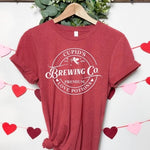 A red tee shirt with cuffed short sleeves. The design is circular with text reading "CUPID'S EST. 1892 BREWING CO. PREMIUM LOVE POTIONS".