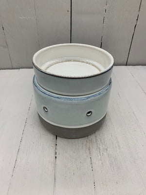 A circular ceramic wax warmer. It is light gray with a band of dark gray on the bottom. There are two holes where light can shine through. The camera shows it from the top down.
