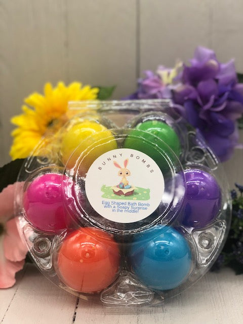 A clear plastic case holding six plastic eggs. They are pink, orange, yellow, green, blue, and purple. It is labeled "Bunny Bombs: Egg Shaped Bath Bomb with a Soapy Surprise in the Middle!"
