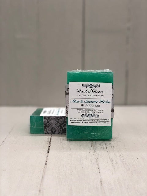 A teal rectangular soap bar. It has a gray damask paper band and a square white label reading "Aloe & Summer Herbs".